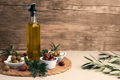 Photo of Bottle of oil, olives and tree twigs on beige table, space for text