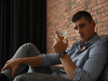 Young man with glass of whiskey near brick wall indoors