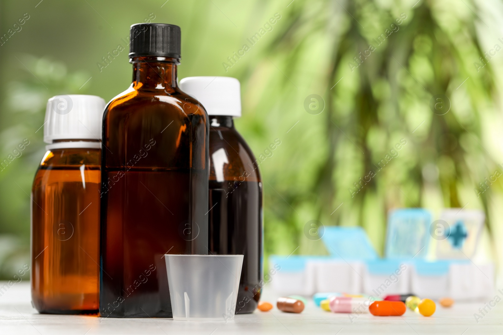 Photo of Bottles of syrup, measuring cup and pills on table against blurred background, space for text. Cold medicine