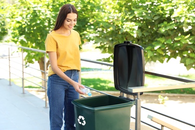 Photo of Young woman throwing plastic bottle into recycling bin outdoors