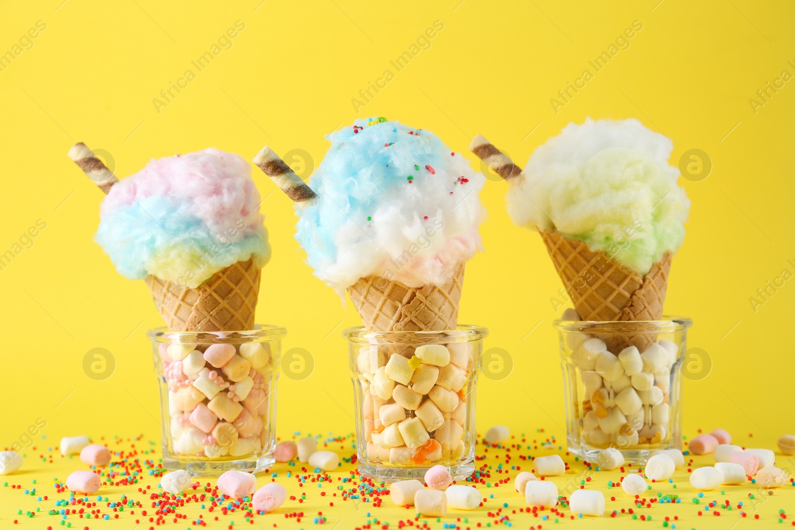 Photo of Sweet cotton candies in waffle cones and marshmallows on yellow background, closeup