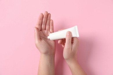 Woman applying cosmetic cream from tube onto her hand on pink background, top view