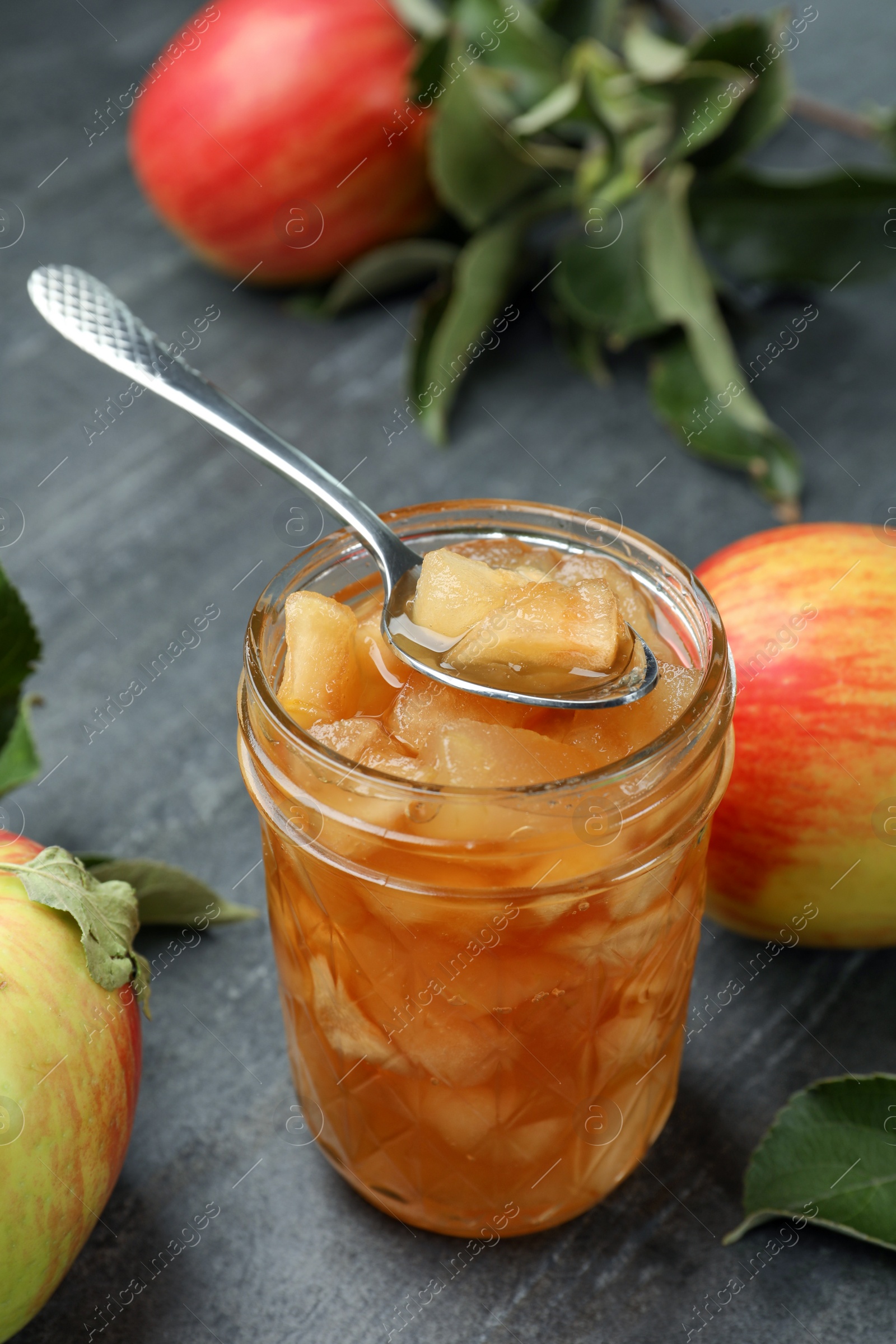 Photo of Tasty apple jam with spoon in glass jar and fresh fruits on grey table