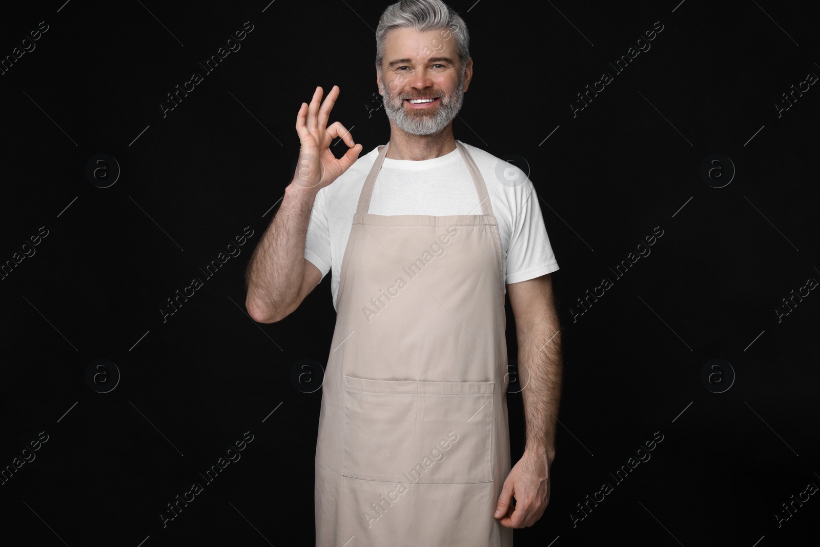 Photo of Happy man in kitchen apron showing OK gesture on black background. Mockup for design