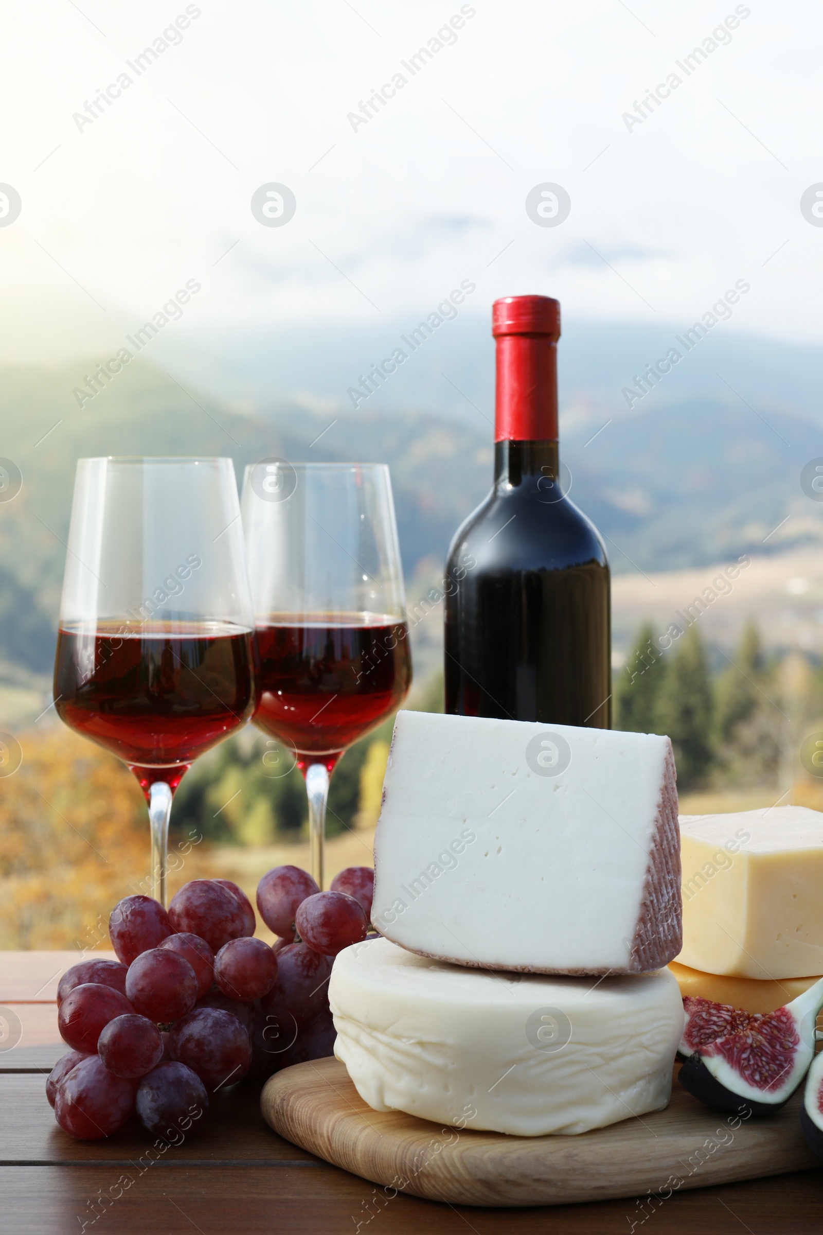 Photo of Different types of delicious cheeses, snacks and wine on wooden table against mountain landscape