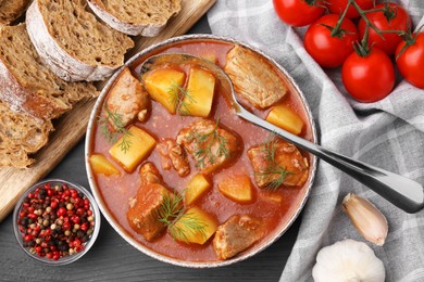 Photo of Delicious goulash in bowl, bread and ingredients on dark wooden table, flat lay