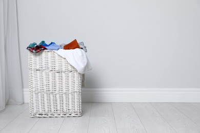 Photo of Wicker laundry basket full of dirty clothes on floor near light wall. Space for text
