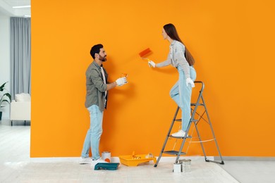 Photo of Man holding can of dye and woman painting orange wall indoors. Interior design