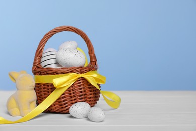 Photo of Wicker basket with festively decorated Easter eggs and bunny on white wooden table against light blue background. Space for text