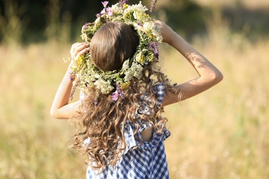 Little girl wearing wreath made of beautiful flowers in field on sunny day