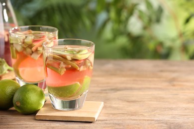 Photo of Glasses of tasty rhubarb cocktail and lime fruits on wooden table outdoors, closeup. Space for text