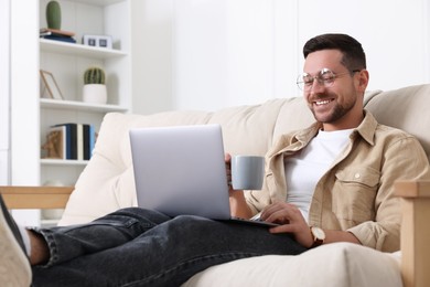 Photo of Happy man with cup of drink working with laptop on sofa at home
