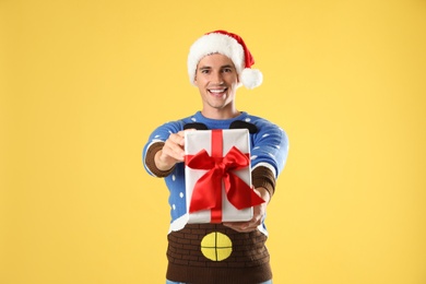 Happy man in Christmas sweater and Santa hat holding gift box on yellow background