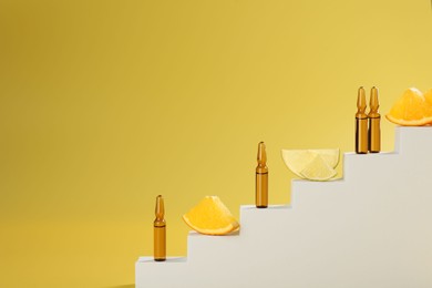 Stylish presentation of skincare ampoules with vitamin C and citrus slices on decorative stairs against yellow background, closeup. Space for text