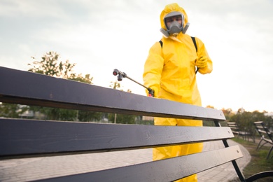 Photo of Person in hazmat suit disinfecting bench in park with sprayer. Surface treatment during coronavirus pandemic