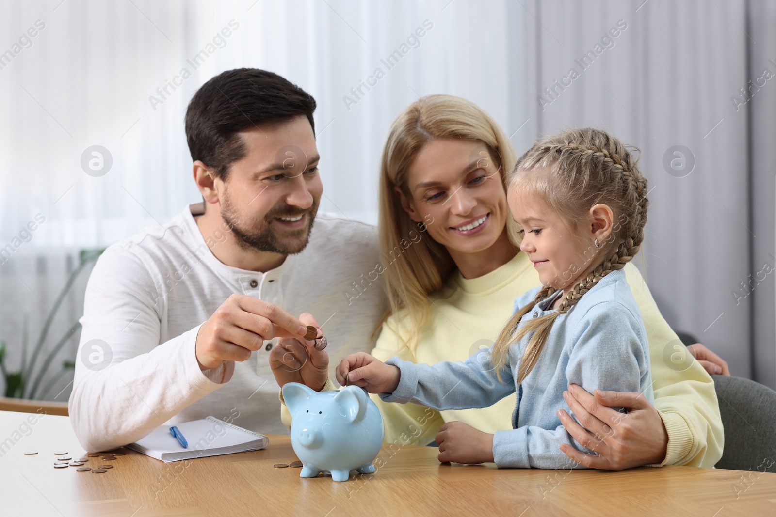 Photo of Planning budget together. Little girl with her family putting coins into piggybank at table indoors