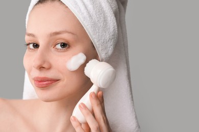 Photo of Young woman washing face with brush and cleansing foam on grey background, closeup. Space for text