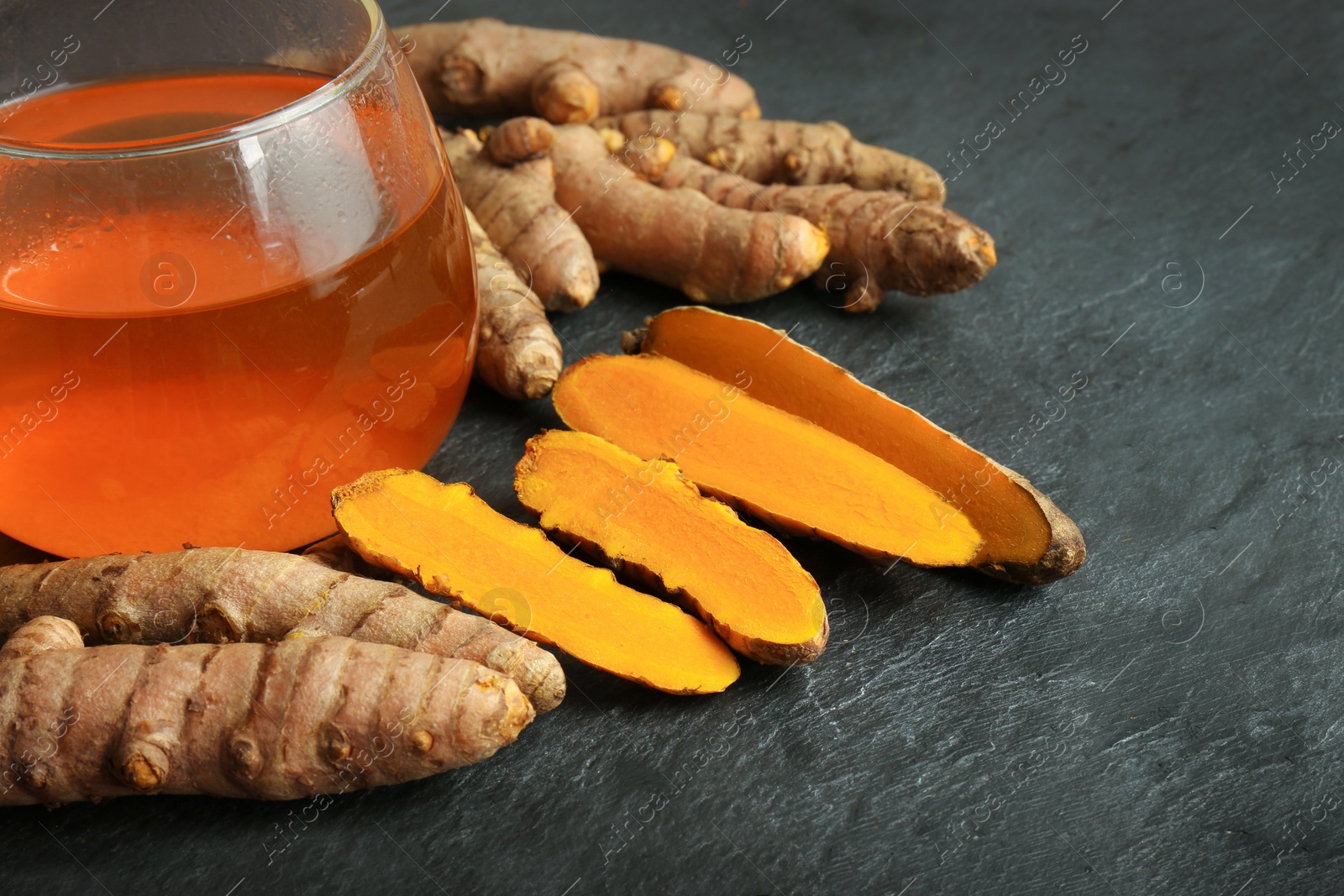 Photo of Glass cup of tasty tea, whole and cut turmeric roots on black textured table, space for text