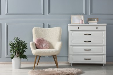 Photo of Stylish armchair with pillow, houseplant and chest drawers near light grey wall indoors. Interior design