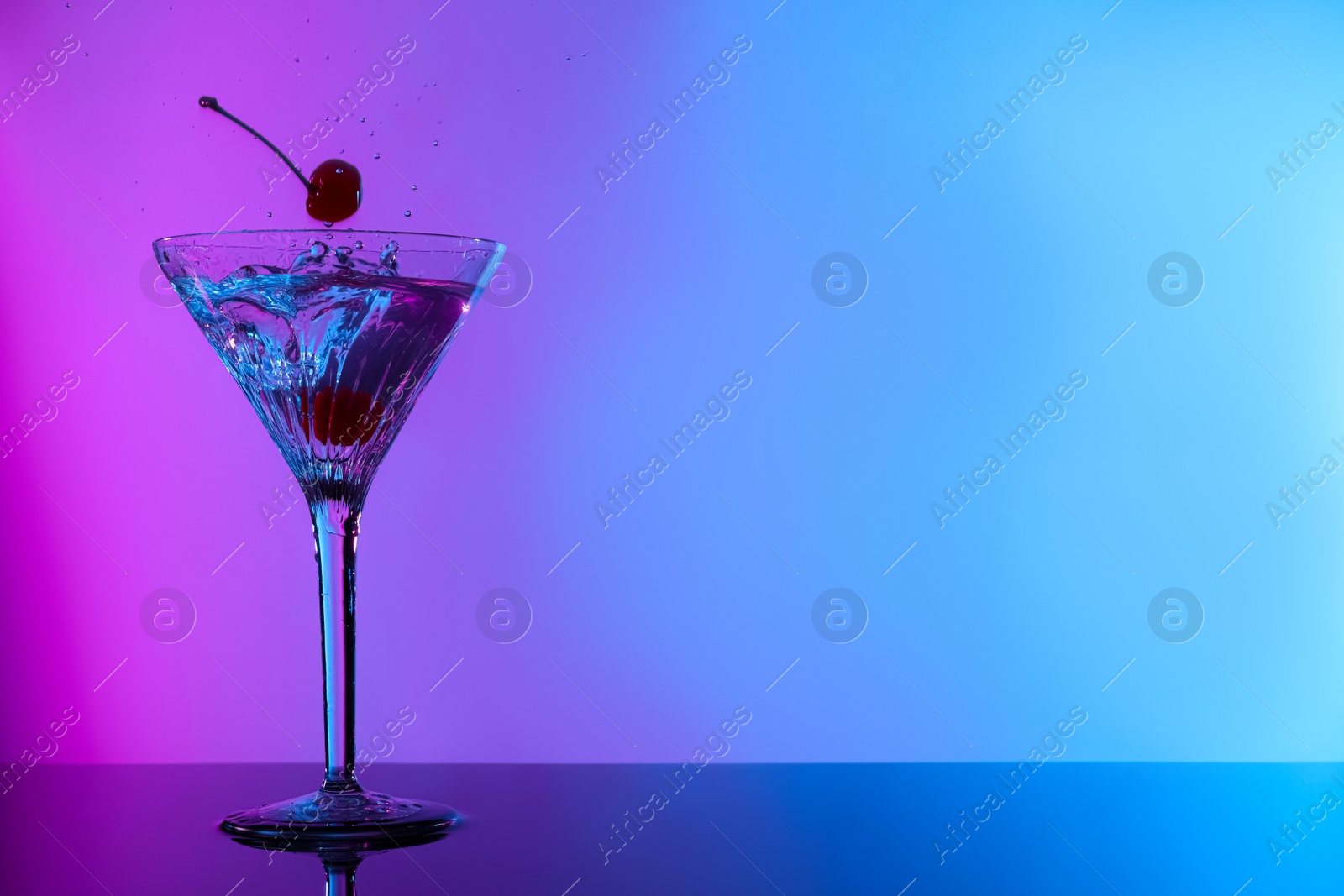Photo of Adding cherries into glass with martini on table in neon lights, space for text