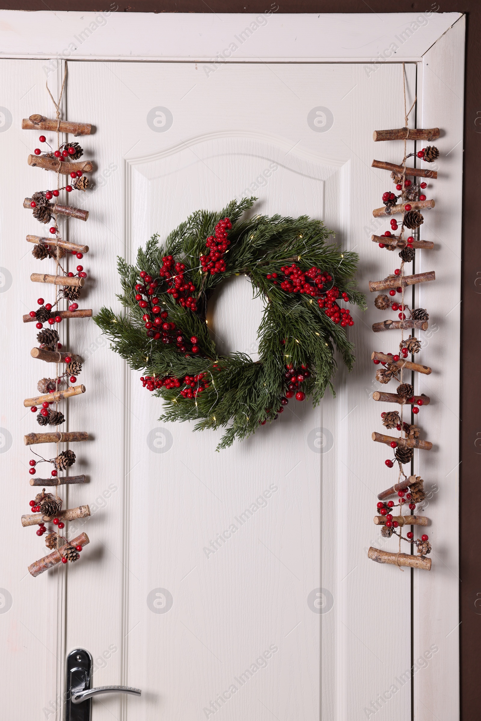 Photo of Beautiful Christmas wreath with red berries and fairy lights hanging on white door