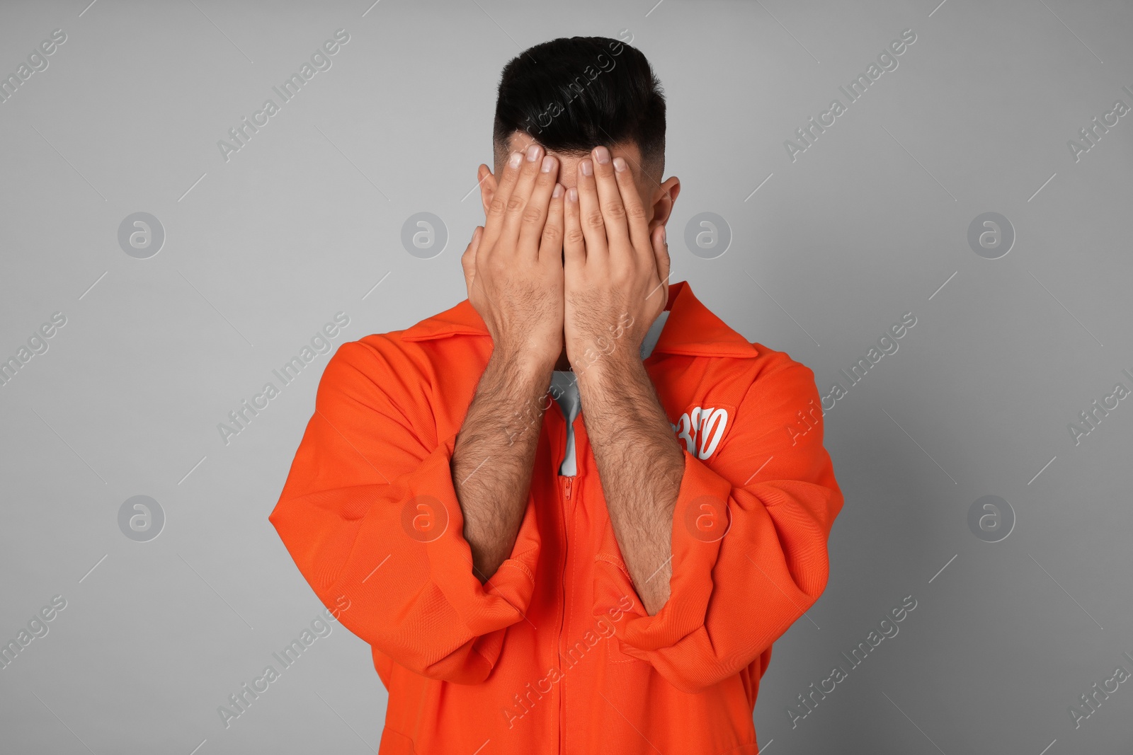 Photo of Remorseful prisoner in orange jumpsuit hiding his face on grey background