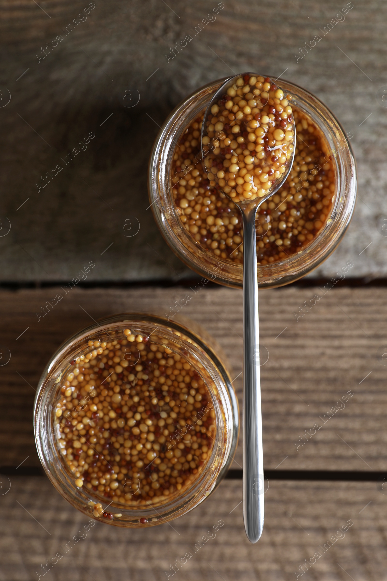 Photo of Jars and spoon of whole grain mustard on wooden table, flat lay