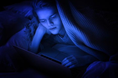 Image of Internet addiction. Teenage boy using laptop under blanket on bed at night. Toned in blue