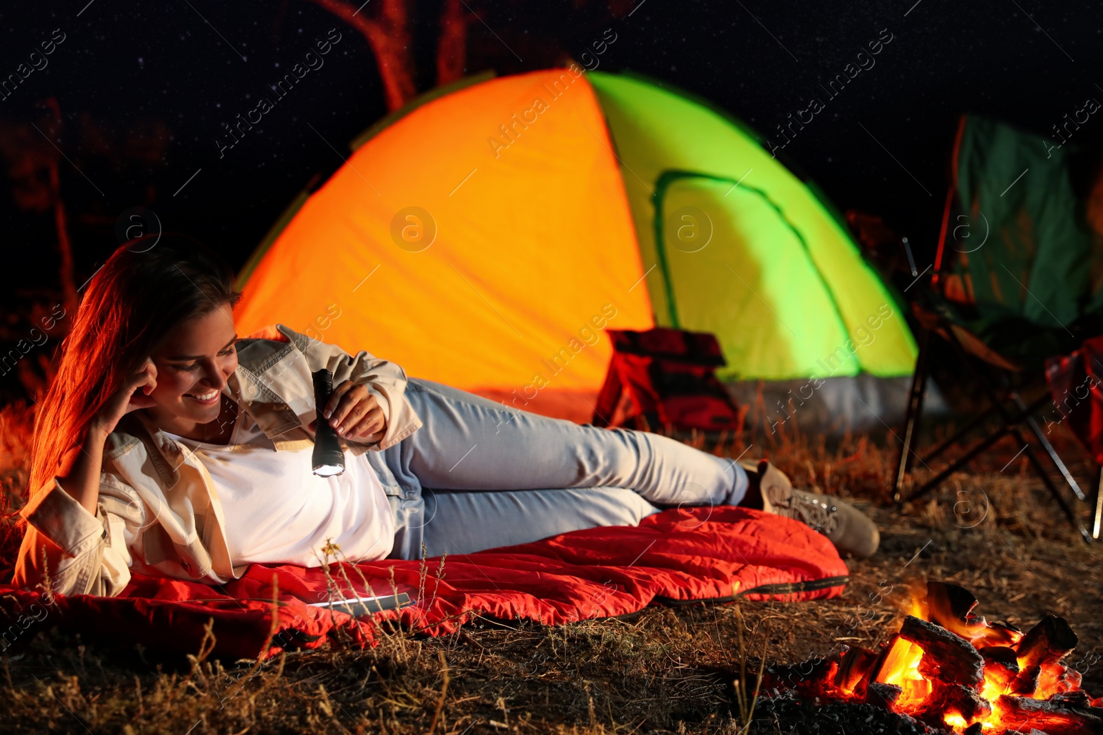 Photo of Young woman with flashlight reading book near bonfire at night. Camping season