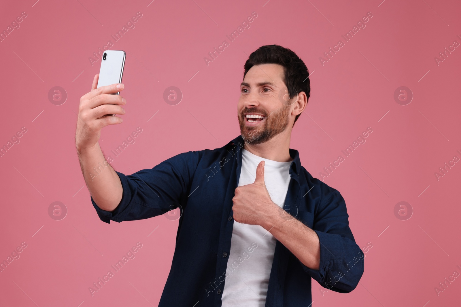 Photo of Smiling man taking selfie with smartphone and showing thumbs up on pink background