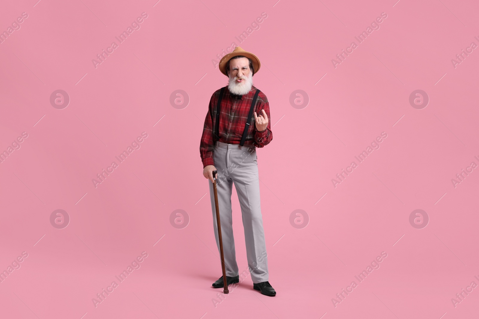 Photo of Senior man with walking cane showing his tongue and rock gesture on pink background