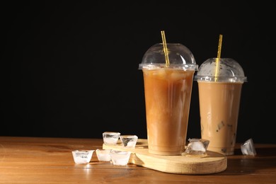 Photo of Refreshing iced coffee with milk in takeaway cups on wooden table against black background, space for text