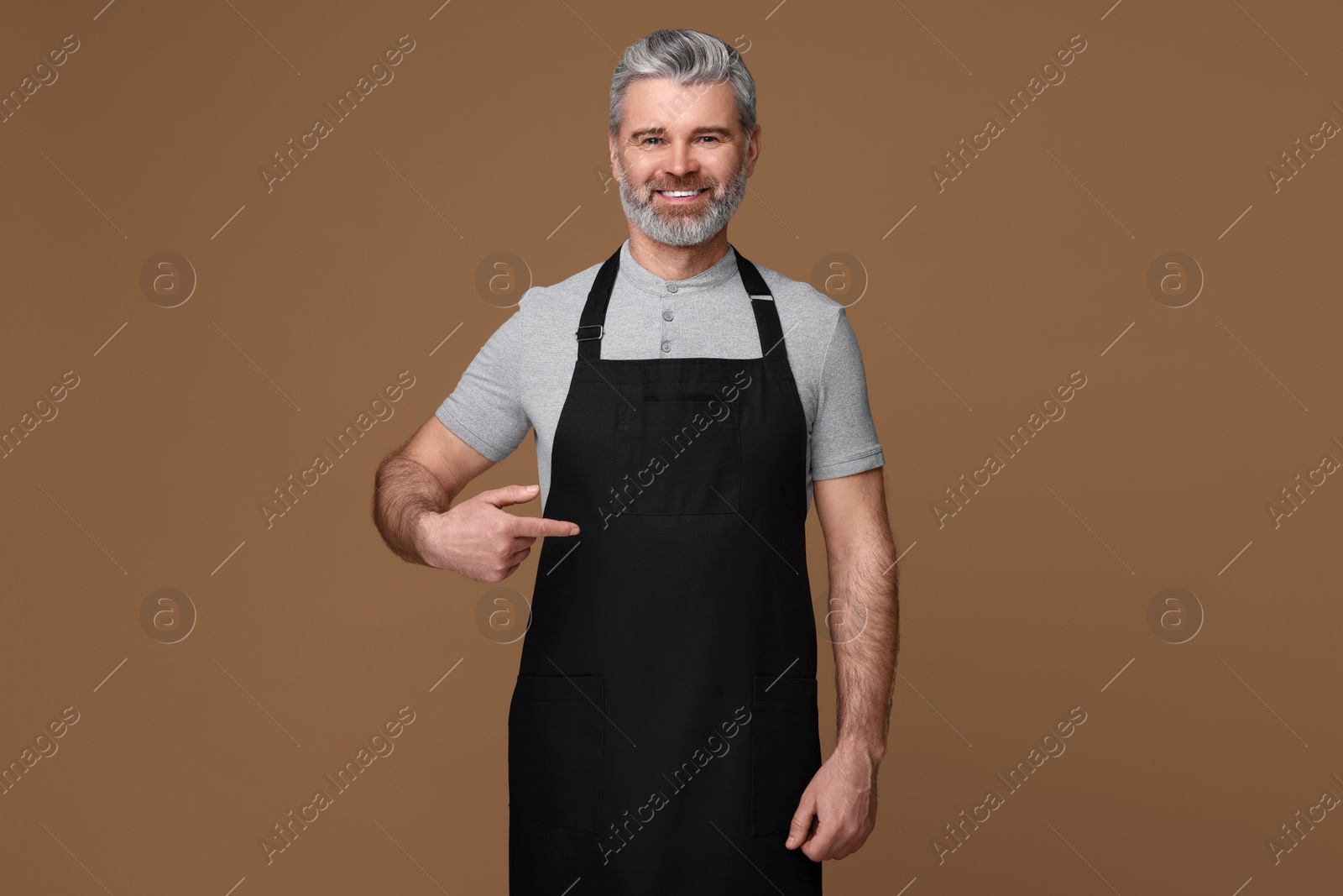 Photo of Happy man pointing at kitchen apron on brown background. Mockup for design