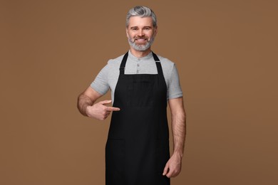 Photo of Happy man pointing at kitchen apron on brown background. Mockup for design