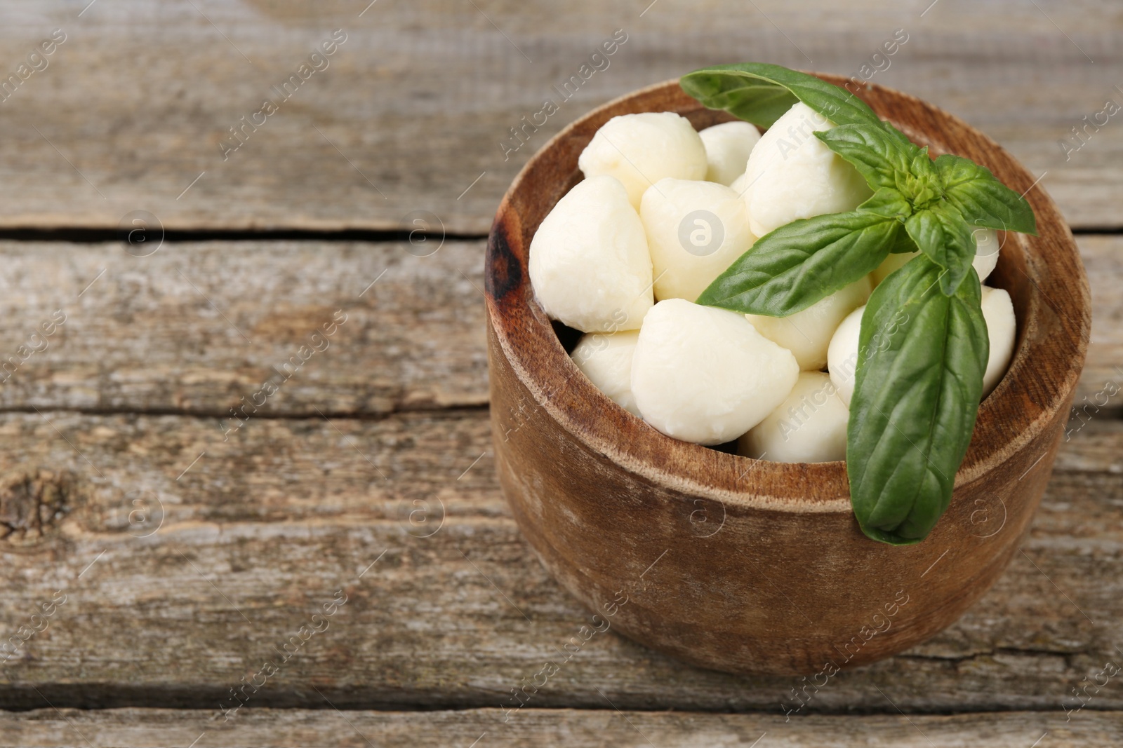 Photo of Tasty mozzarella balls and basil leaves in bowl on wooden table, closeup. Space for text