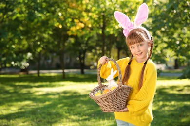 Photo of Cute little girl with bunny ears and basket of Easter eggs in park. Space for text