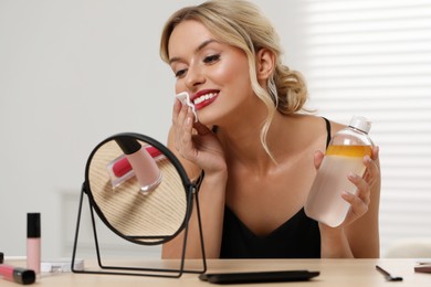 Photo of Smiling woman removing makeup with cotton pad in front of mirror at table indoors