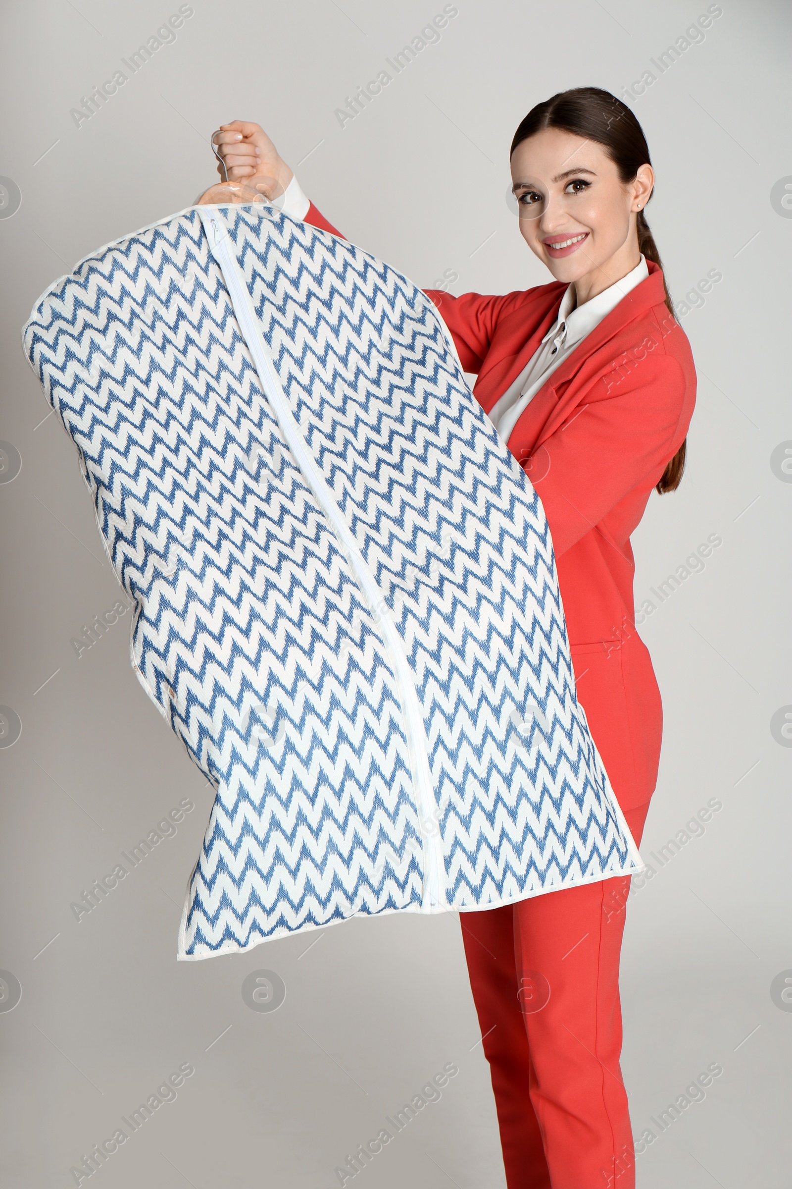 Photo of Young woman holding hanger with clothes in garment cover on light grey background. Dry-cleaning service
