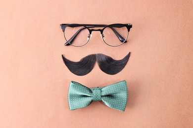 Man's face made of artificial mustache, glasses and bow tie on brown background, top view