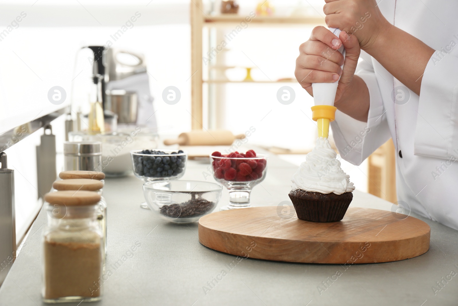 Photo of Young female pastry chef decorating cupcake with cream at table in kitchen, closeup