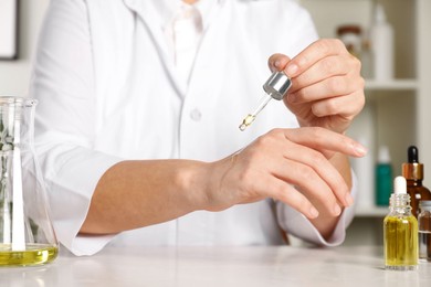 Photo of Dermatologist testing cosmetic oil at white table indoors, closeup