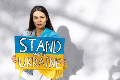 Photo of Young woman holding poster in colors of national flag and words Stand with Ukraine near light wall. Space for text