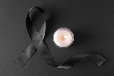 Photo of Black ribbon and burning candle on dark background, top view. Funeral symbols