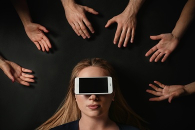 Young woman with smartphone covering her eyes surrouded by people's hands on black background, top view. Space for text