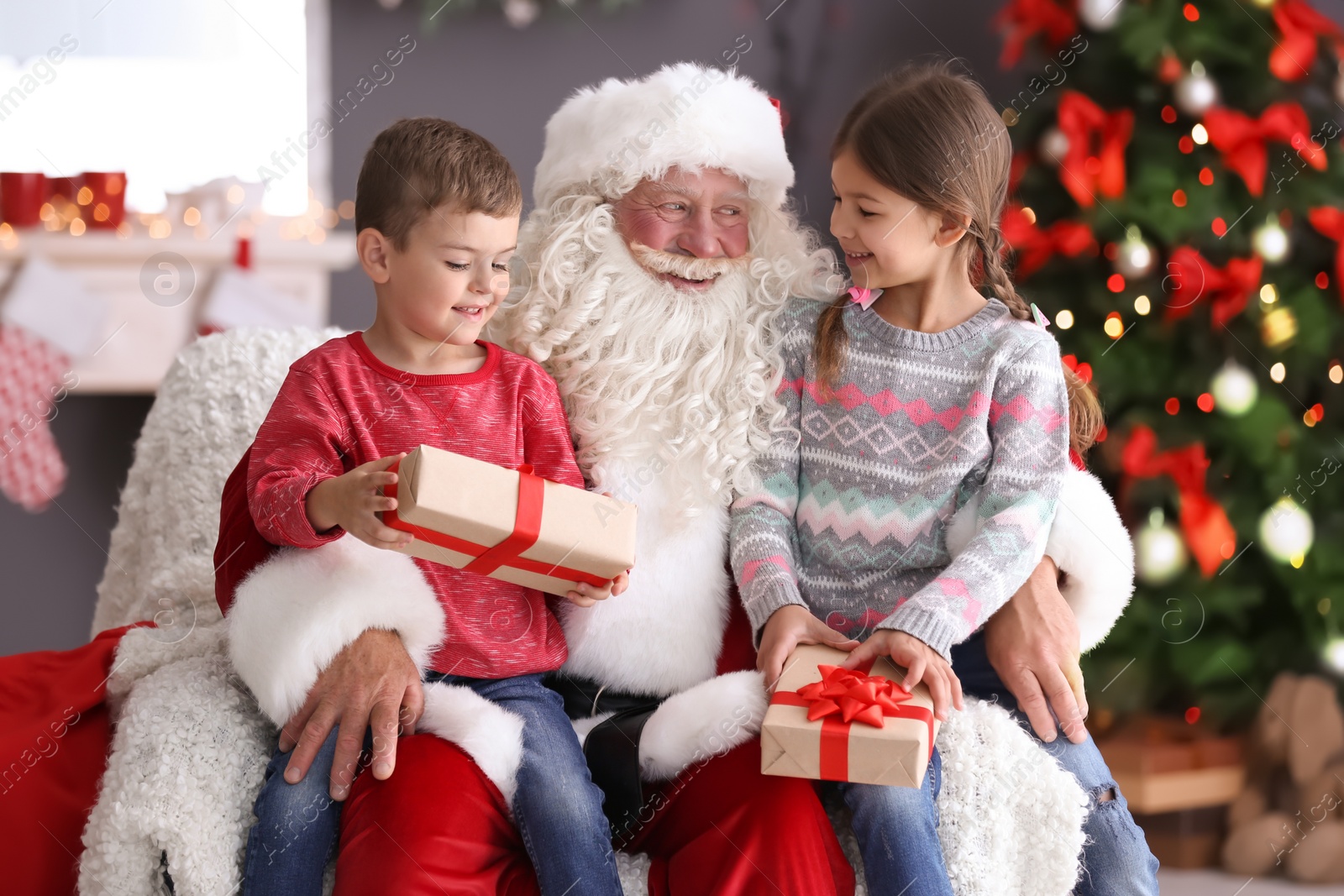 Photo of Little children with gift boxes sitting on authentic Santa Claus' knees indoors