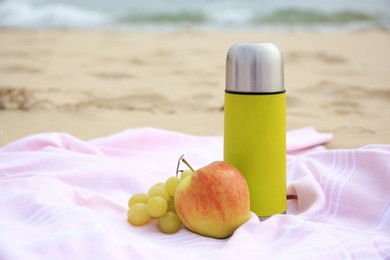 Metallic thermos with hot drink, fruits and plaid on sandy beach near sea