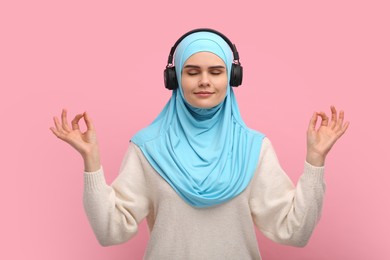 Photo of Muslim woman in hijab and headphones meditating on pink background