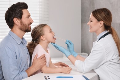 Photo of Smiling doctor examining girl`s oral cavity with tongue depressor near her father indoors