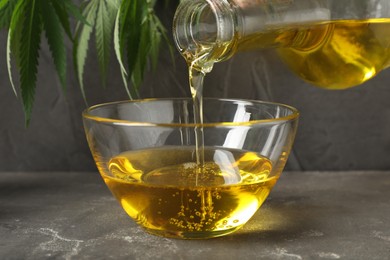 Photo of Pouring hemp oil into glass bowl on grey table, closeup