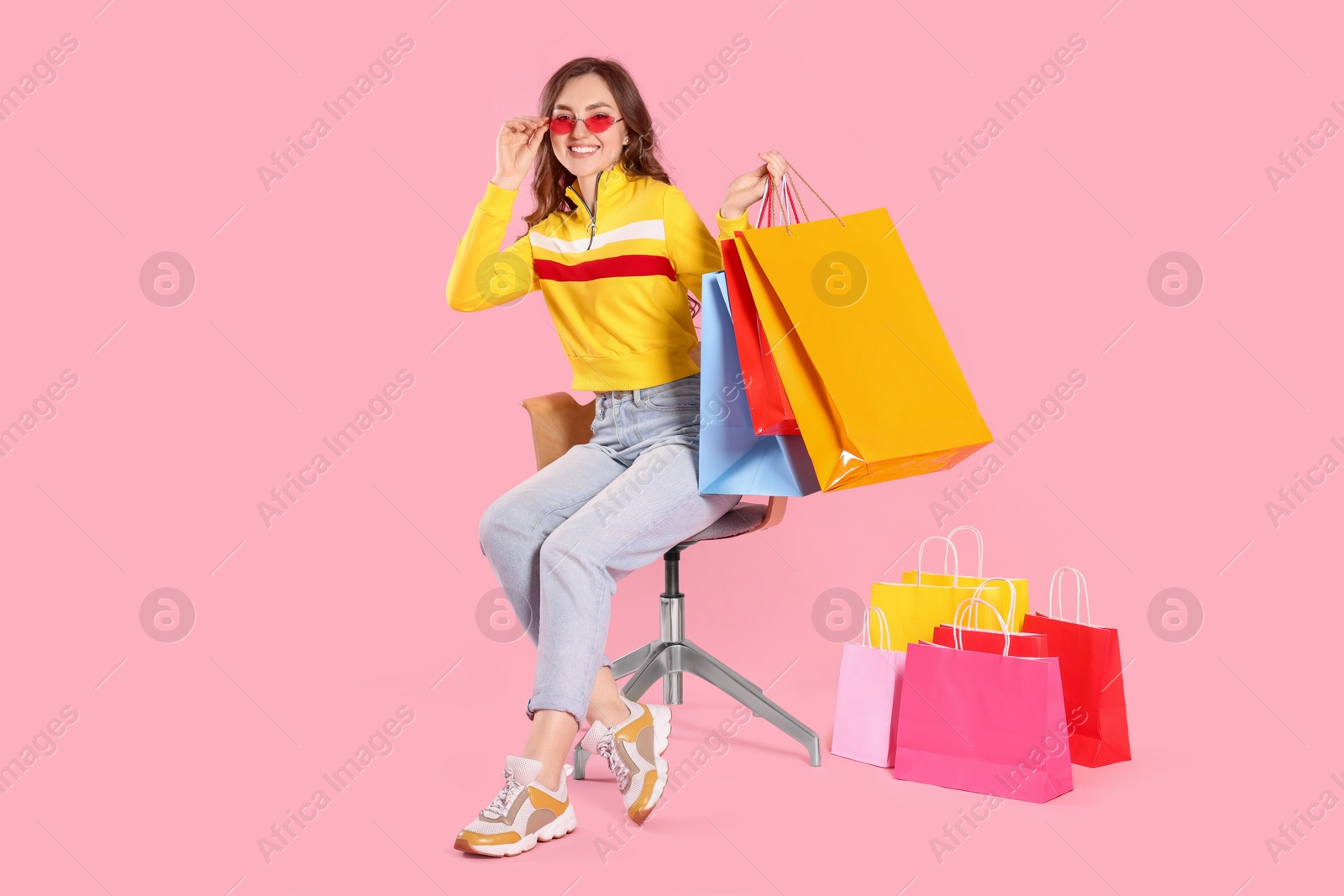 Happy woman in stylish sunglasses holding many colorful shopping bags on armchair against pink background Photo of Happy woman in stylish sunglasses holding many colorful shopping bags on armchair against pink background
